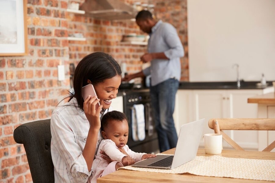 A woman with a baby on the phone using a laptop at a breakfast table, while her husband is at the stove in the background.