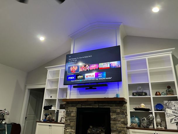 A mounted TV with glowing blue backlighting above a stone fireplace, surrounded by white built-in shelves.