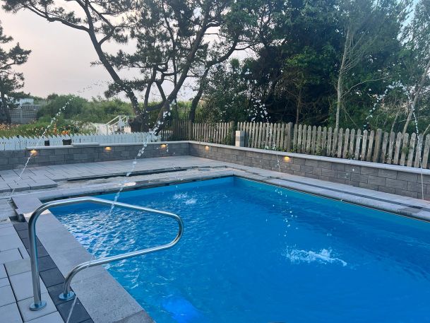 A backyard swimming pool with fountains and a paved area featuring lounge chairs and a wooden fence.