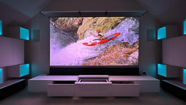 A home theater setup with a large projector screen showing a kayaker on a waterfall, surrounded by illuminated shelves.