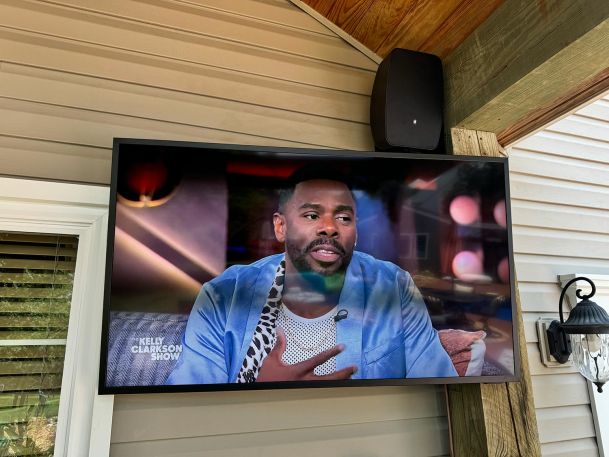 Outdoor TV mounted under a covered patio playing a daytime talk show.