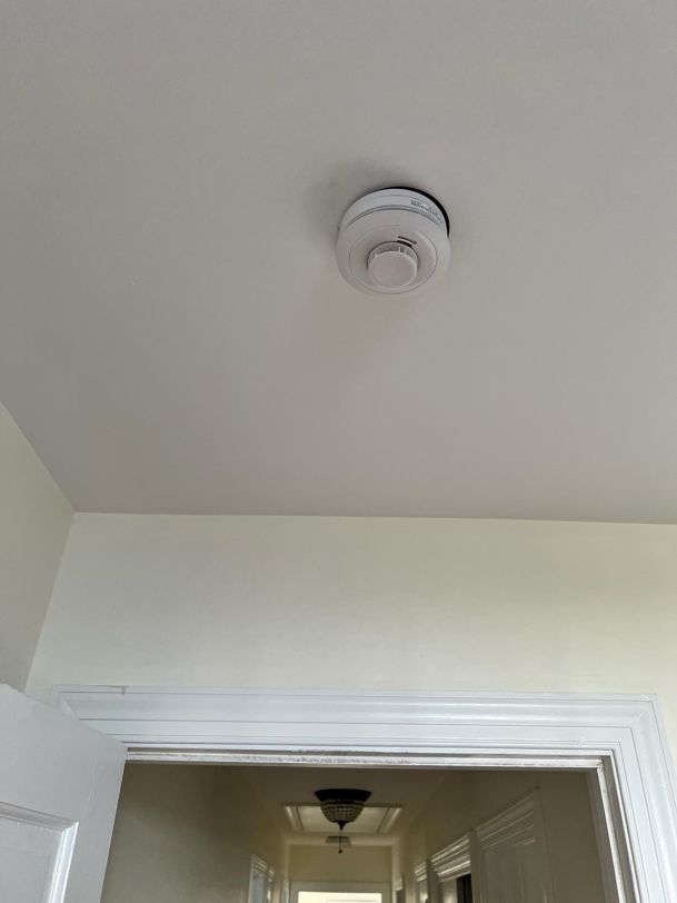 A hallway with a smoke detector and attic access door in the ceiling, with simple beige walls.