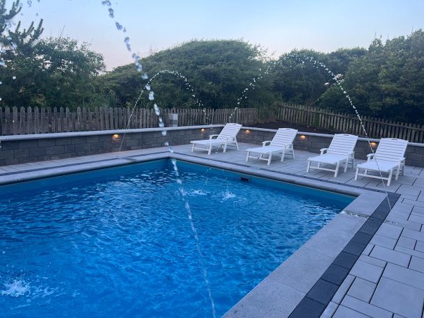 A closer view of a backyard swimming pool, showcasing deck fountains and surrounding greenery.