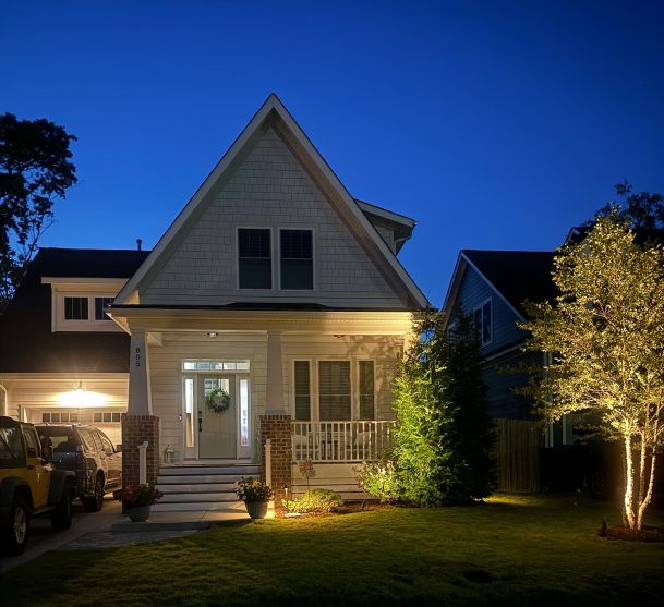 A cozy house at dusk with soft exterior lighting highlighting the front porch and trees.