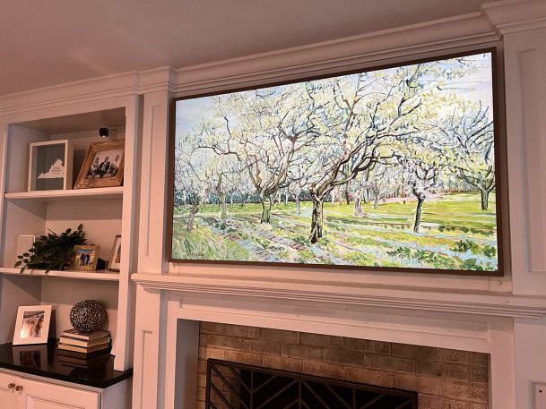 A mounted TV above a fireplace, framed as an art piece displaying a painting of a spring orchard.