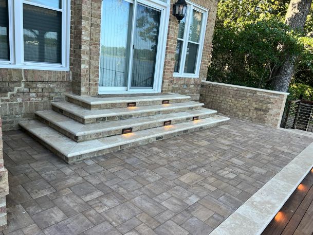 A paved outdoor patio with brick steps illuminated by step lights, leading to a house with large windows.