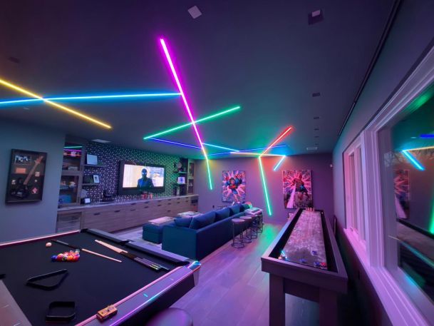 A colorful game room featuring neon ceiling lights, a pool table, and a shuffleboard table.