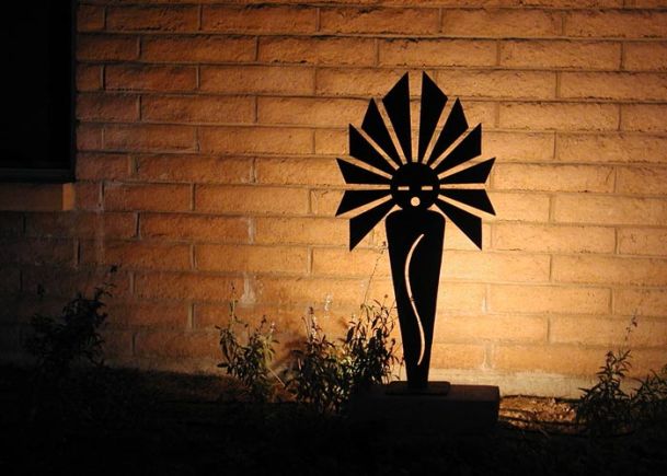 A silhouetted metal sculpture illuminated against a brick wall in an outdoor garden setting.