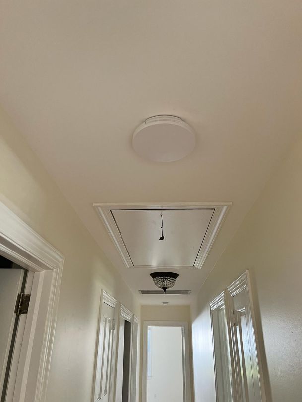 A hallway with a smoke detector and attic access door in the ceiling, with simple beige walls.